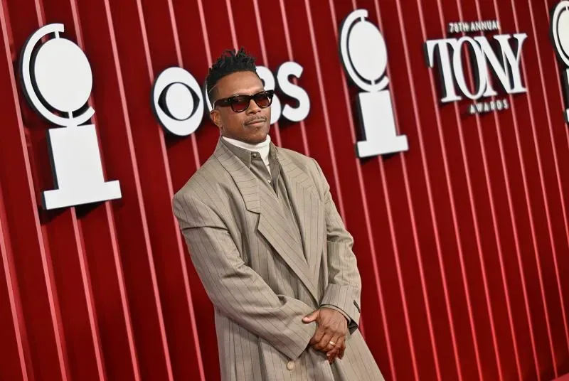 أحد نجوم حفل توزيع جوائز توني الـ78، يرتدي بدلة رمادية عصرية ونظارات شمسية، يقف على السجادة الحمراء أمام لافتة جوائز توني.