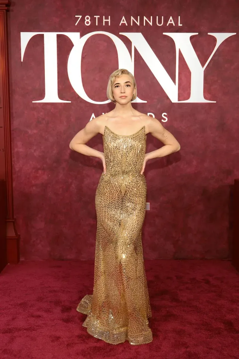 أريانا ديبوس تتألق بفستان ذهبي لامع في حفل توزيع جوائز توني الـ78، مع خلفية حمراء تعكس أجواء الحدث الفاخرة.