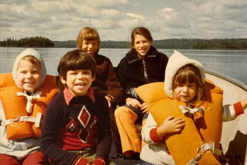 مجموعة من الأطفال يرتدون سترات نجاة، يجلسون في قارب على بحيرة، مع مناظر طبيعية خلابة في الخلفية، تعكس لحظات من الطفولة البسيطة.