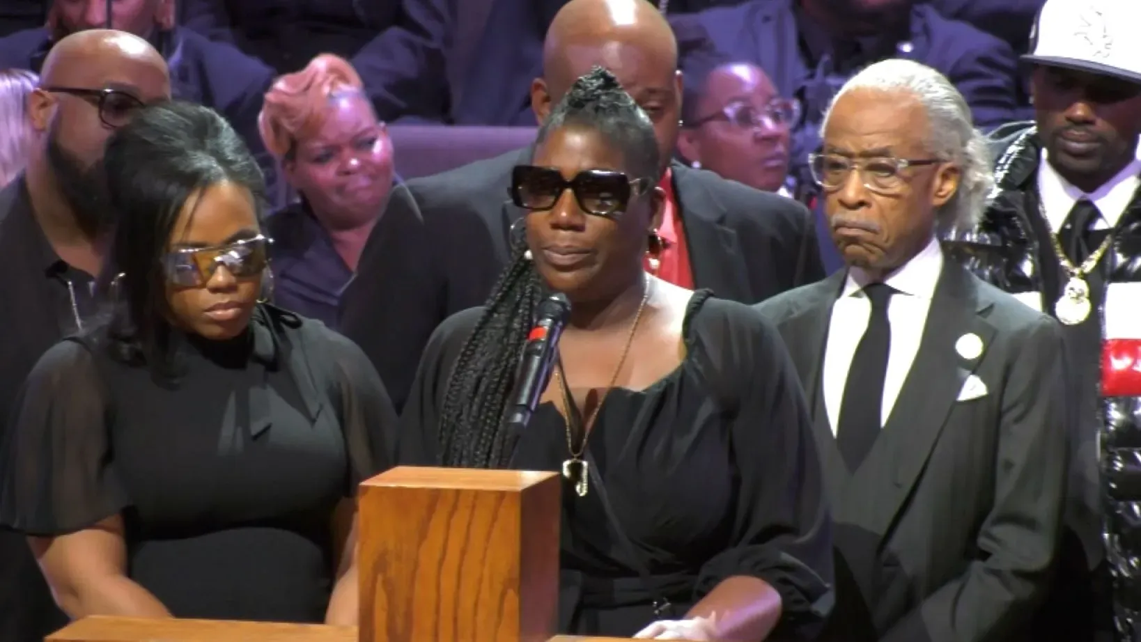 امرأة ترتدي نظارات شمسية تتحدث إلى الحضور في حدث تأبيني، بينما يقف بجانبها شخصان آخران. يبدو التأثر واضحًا على وجوه الحضور.