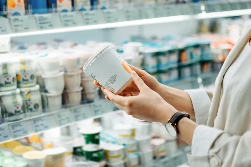 امرأة تقف أمام رف من الزبادي، تحمل علبة زبادي وتفحص المعلومات الغذائية، تعكس مشاعر القلق بشأن المحليات الصناعية.