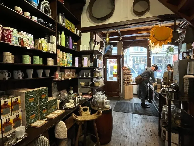 داخل متجر "t Zonnetje" في أمستردام، يظهر زبونان يتسوقان بين أرفف مليئة بالشاي والتوابل، مع تفاصيل تاريخية مميزة.