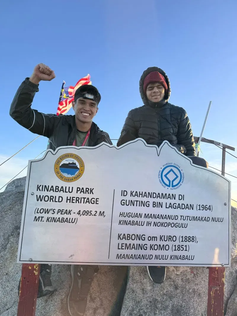 صورة لاثنين من المتسلقين عند قمة جبل كينابالو، يحتفلان ببلوغ القمة أمام لافتة تشير إلى ارتفاع الجبل وتاريخ المنطقة.