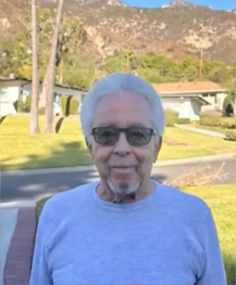 رجل مسن يرتدي نظارات شمسية وقميص رمادي، يقف في حديقة أمام منازل، مع جبال في الخلفية، في سياق حرائق لوس أنجلوس.