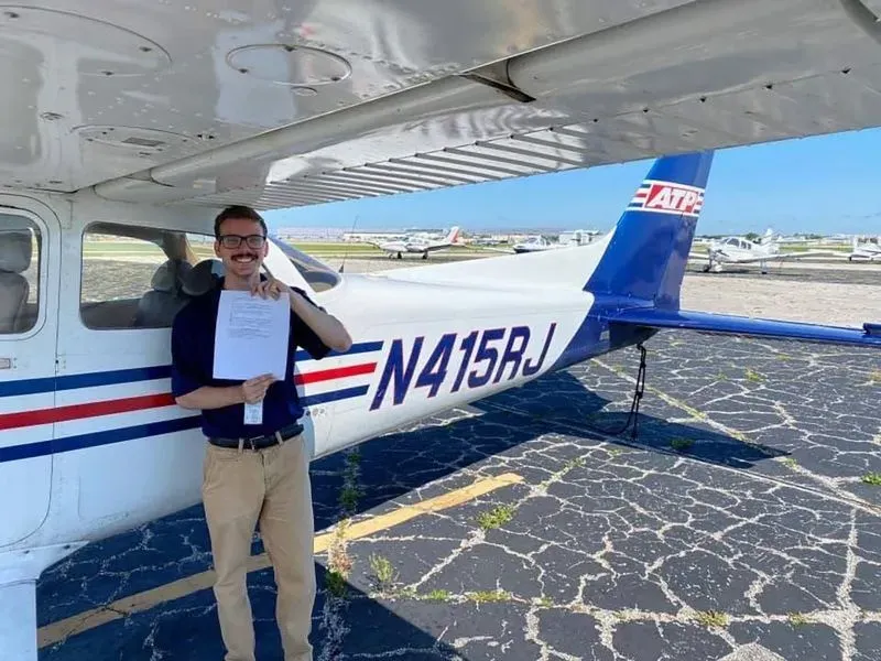 شاب مبتسم يقف بجانب طائرة صغيرة من طراز Cessna، ممسكًا بشهادة. الصورة تعكس شغفه بالطيران وتحقيقه لطموحاته المهنية.