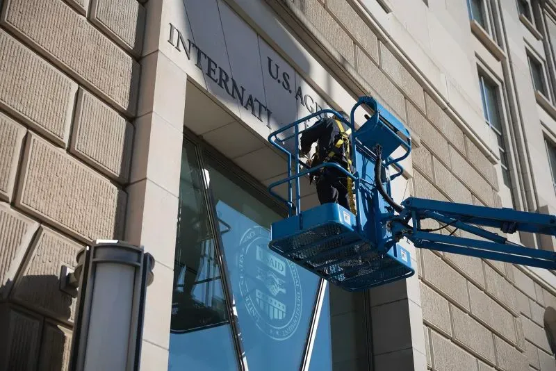 عامل يقوم بتعديل لافتة على واجهة مبنى الوكالة الأمريكية للتنمية الدولية، مع التركيز على التحديات التي يواجهها برنامج "ذا إنفوي شو".