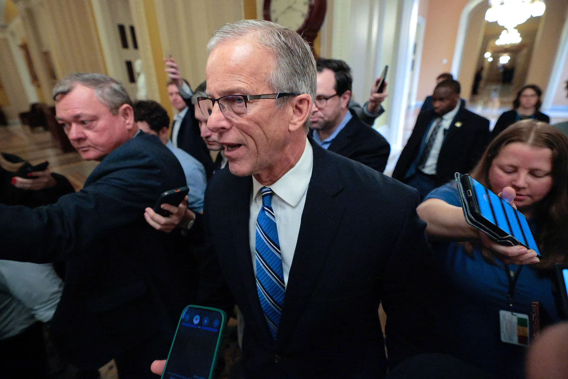 مجموعة من الأشخاص في مبنى حكومي، مع التركيز على رجل يرتدي بدلة وقميصًا أزرق، يتحدث مع الصحفيين حول تمويل وزارة الأمن الداخلي.