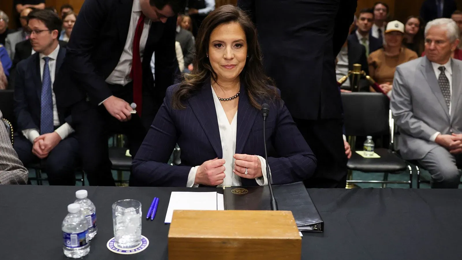 النائبة إليز ستيفانيك تجلس أمام طاولة في جلسة استماع، مع زجاجات مياه وملاحظات، وسط حضور عدد من الأشخاص في الخلفية.