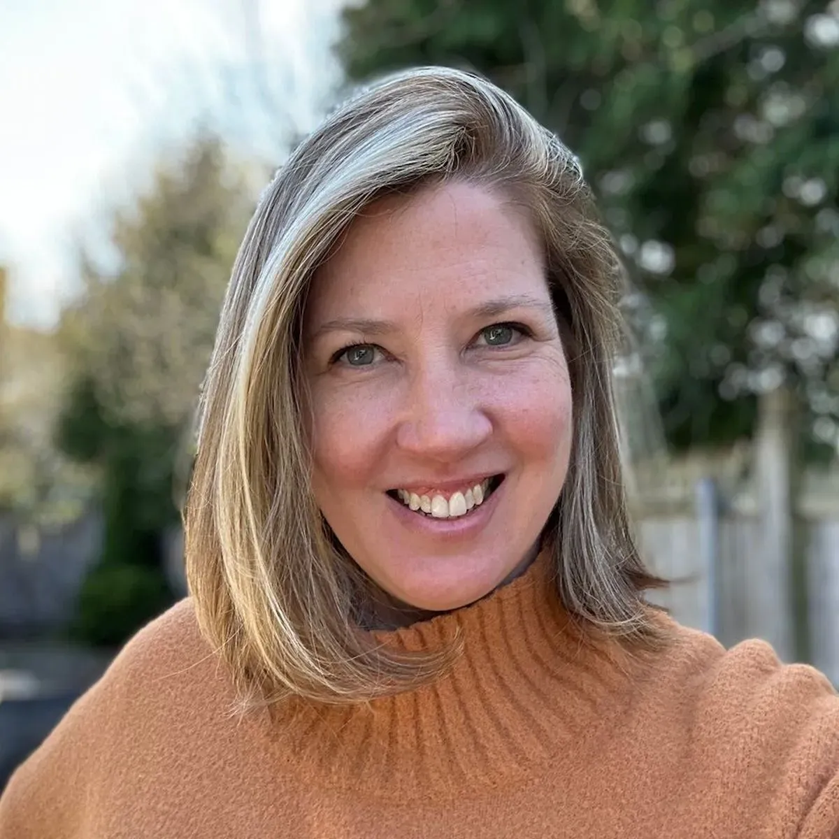 امرأة مبتسمة ترتدي سترة برتقالية، تظهر في خلفية طبيعية خضراء، تعكس الأمل والمرونة بعد حادث مأساوي.