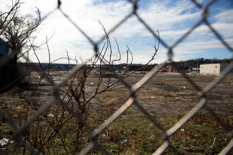 منظر لقطعة أرض مهجورة محاطة بسياج، يظهر فيها بعض الأعشاب الجافة والنفايات، مع سماء زرقاء وصافية في الخلفية.