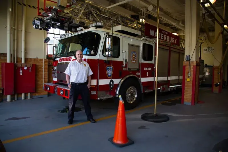 رجل يرتدي زي الإطفاء يقف أمام شاحنة إطفاء في محطة إطفاء واتربري، مع تفاصيل حول الحادثة المأساوية في المنطقة.