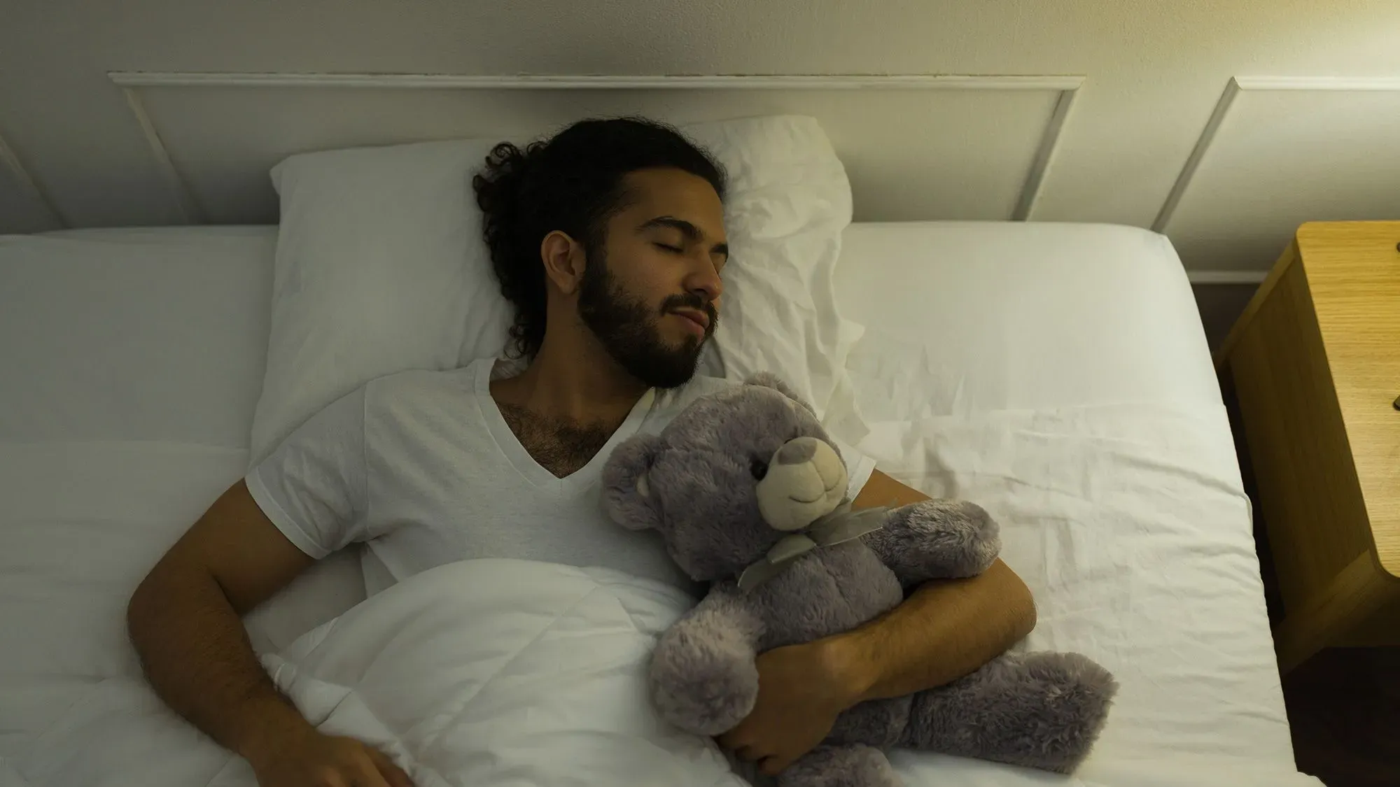 رجل نائم في سريره يحتضن دمية دب رمادية، مما يعكس أهمية الحيوانات المحشوة في توفير الراحة والهدوء للبالغين.