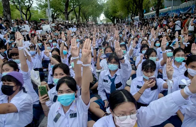 طلاب يرتدون زيًا موحدًا يجلسون في تجمع احتجاجي، رافعين أيديهم، تعبيرًا عن المطالب بتخفيف قيود تسريحات الشعر في تايلاند.