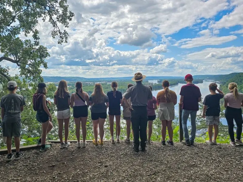 مجموعة من الطلاب يقفون مع معلمهم في حديقة طبيعية، يتطلعون إلى منظر خلاب للنهر وتلال إفيجي تحت سماء مليئة بالغيوم.