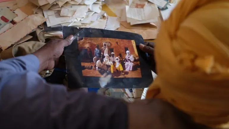 صورة لرجل يحمل صورة قديمة لمجموعة من الأشخاص في أغاديز، محاطًا بأوراق مكدسة، تعكس تاريخ المدينة الثقافي.