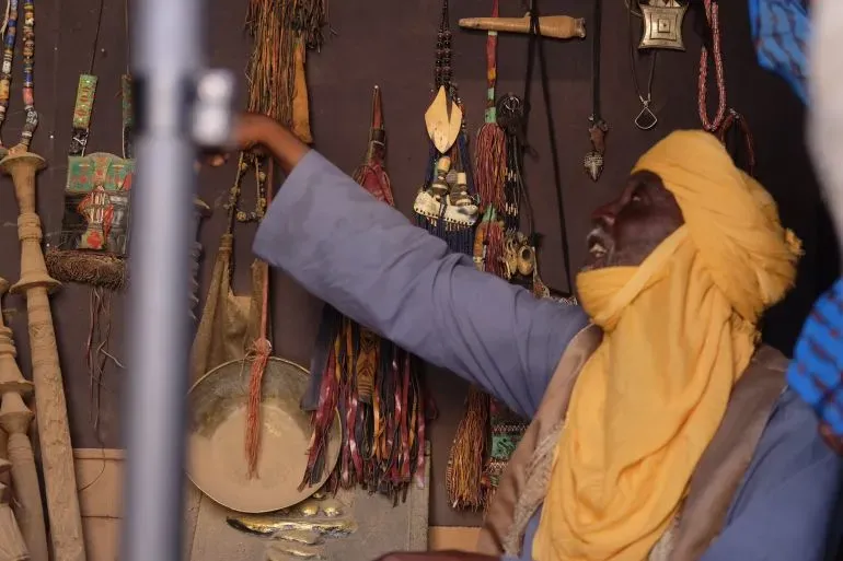 الحاج أمادو ديزي إيلو في متجره لبيع التذكارات بأغاديز، محاطاً بمجوهرات وسكاكين تقليدية، يعكس تراث المدينة الثقافي.