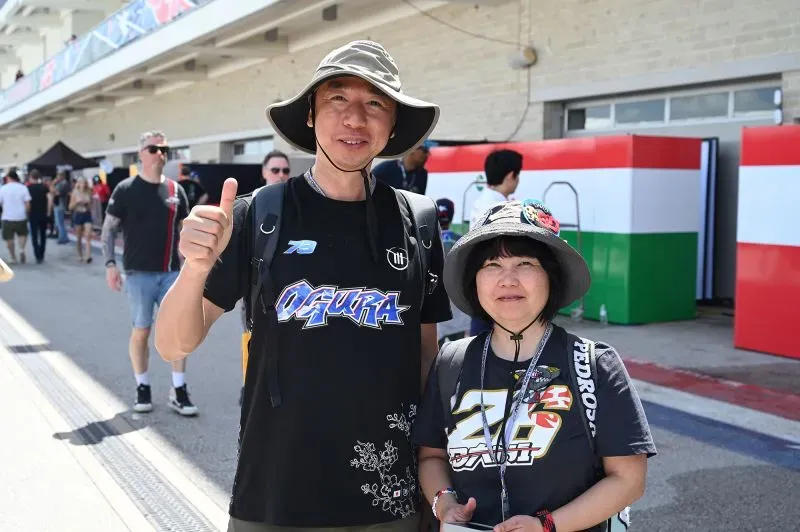 آي أوغورا، متسابق موتو جي بي، يقف مبتسمًا بجوار شقيقته كارين، في حلبة سباق، مرتديًا قمصان رياضية وقبعات.