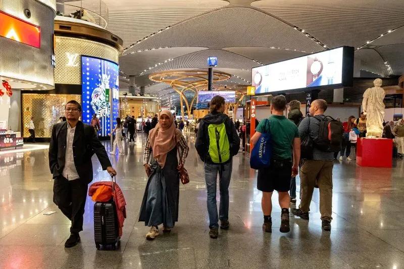 مطار حديث مزدحم بالمسافرين، يظهر تنوعًا ثقافيًا في الأزياء والأمتعة، مع لافتات تجارية وإضاءة مميزة.