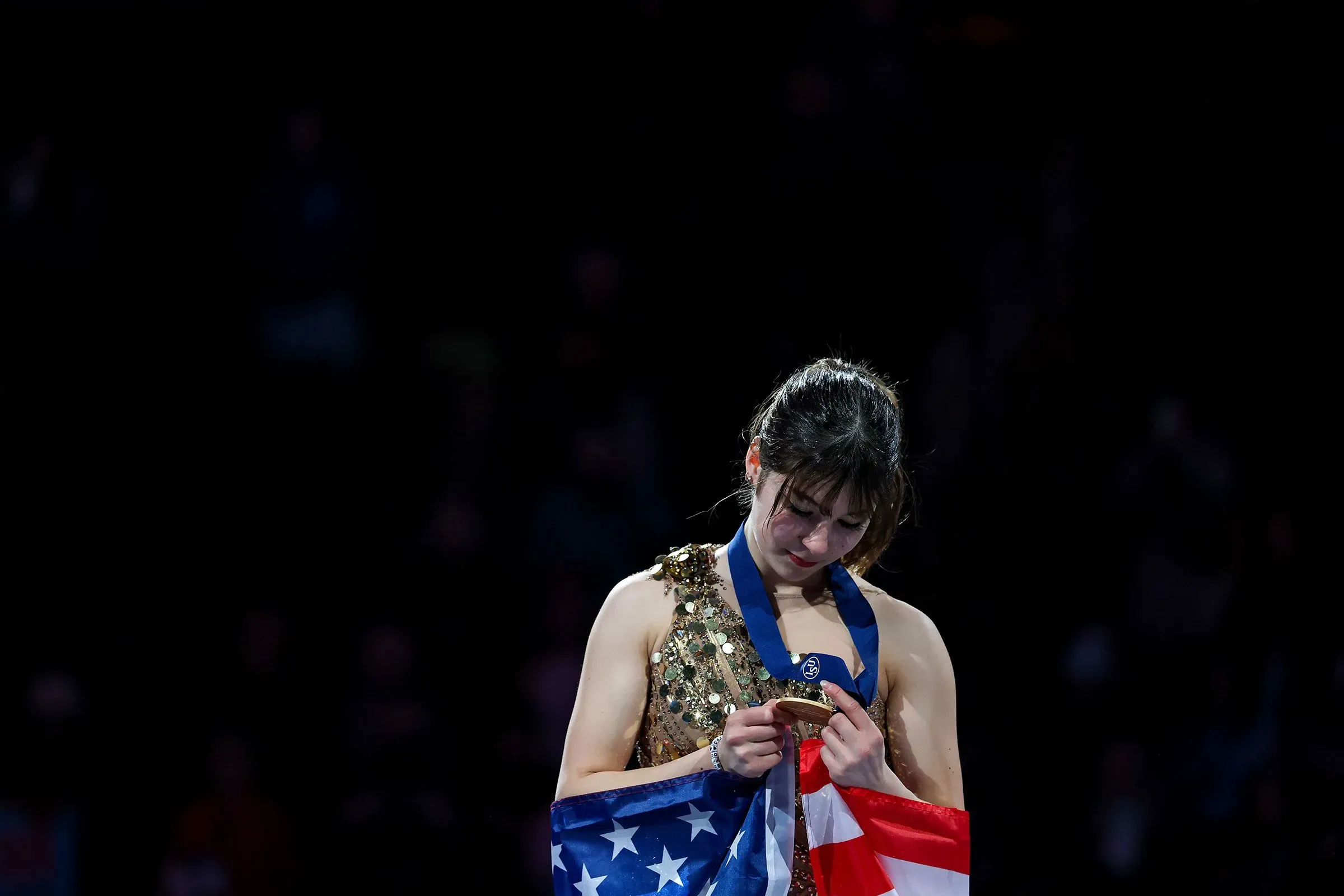 أليسا ليو تحمل ميدالية برونزية بينما تنظر إلى أسفل بتأمل، مرتدية زيها الرياضي، مع العلم الأمريكي خلفها في أجواء احتفالية.