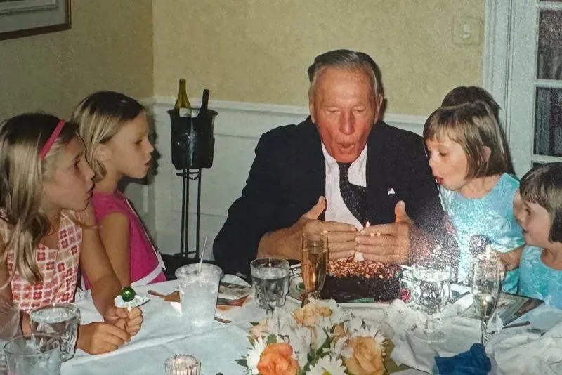 رجل مسن يحتفل بعيد ميلاده مع أربع فتيات صغيرات حول طاولة، حيث يظهرون في لحظة مميزة أثناء إطفاء الشموع.