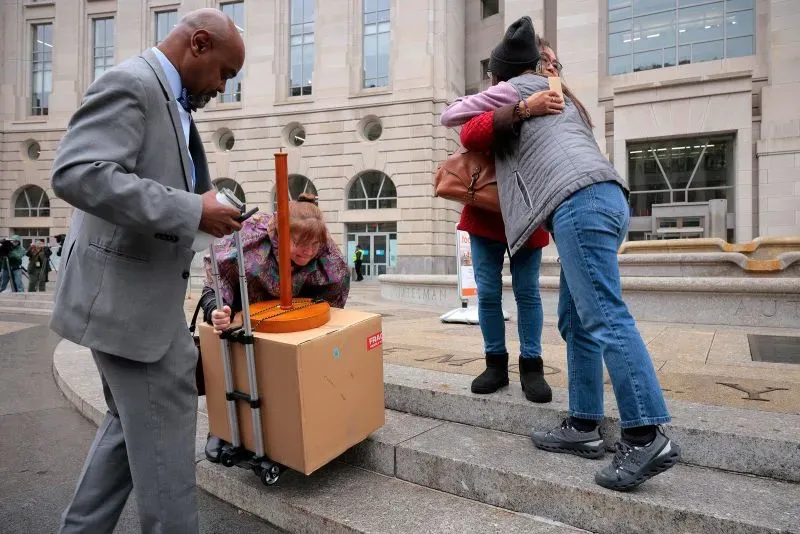 امرأة تحمل صندوقاً كبيراً على عربة، بينما تحتضنها امرأتان أخريتان في مشهد مؤثر أمام مبنى حكومي.