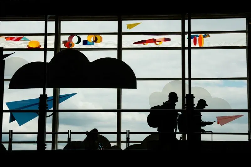 ظلّ silhouetted figures of travelers walk past large windows adorned with colorful paper airplane decorations, reflecting a busy airport atmosphere.