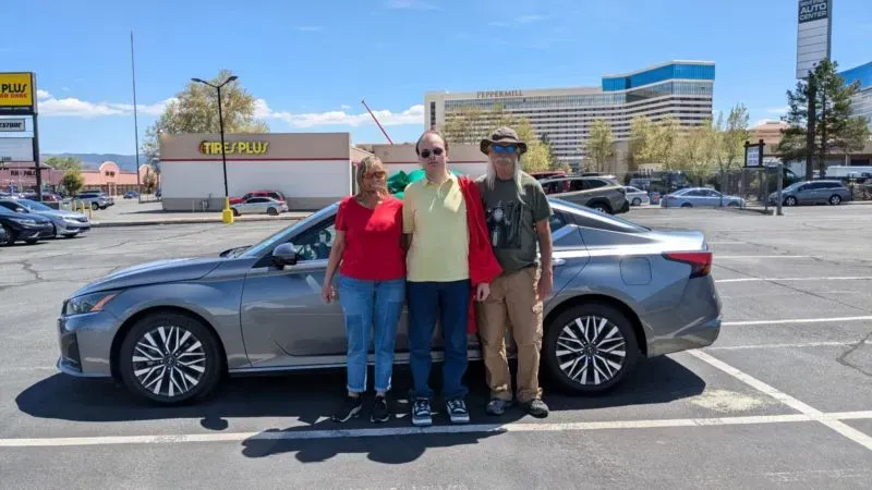 عائلة تتكون من ثلاثة أفراد تقف أمام سيارة جديدة في موقف سيارات، تعكس التحديات المالية بسبب التضخم وارتفاع الأسعار.