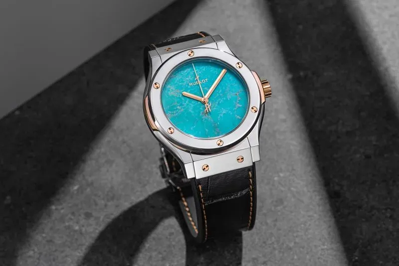 ساعة من هوبلو بتصميم أنيق، ميناء من الفيروز، وحزام جلدي أسود، تظهر في إضاءة طبيعية، تعكس الفخامة والأناقة.