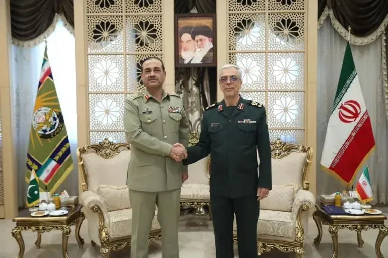 القائد العسكري الباكستاني عاصم منير يتصافح مع نظيره الإيراني في اجتماع رسمي، مع العلم الوطني لكلا البلدين في الخلفية.