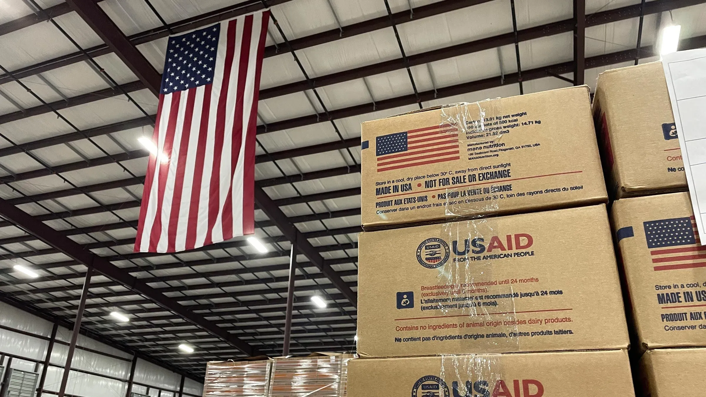 صناديق تحمل شعار USAID مكدسة في مستودع، مع العلم الأمريكي معلق في الأعلى، تمثل جهود الإغاثة لمواجهة سوء التغذية.