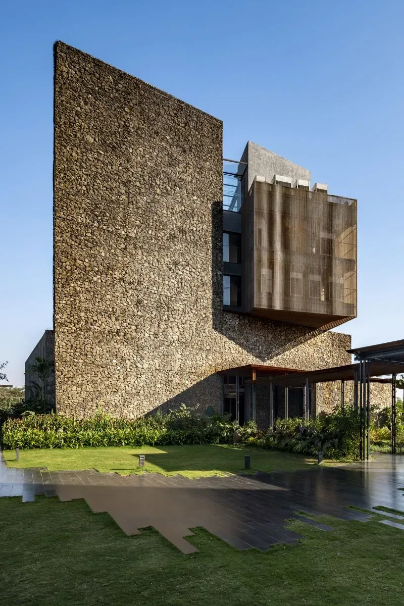 تصميم معماري حديث يظهر جدران حجرية وأسطح خشبية، مع حديقة خضراء في المقدمة، يعكس مفهوم بناء مستدام باستخدام الحمم البركانية.