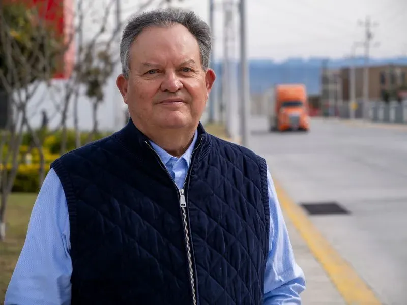 رجل مسن يقف مبتسمًا بخلفية تحتوي على مصانع وشاحنة، يعكس التحول الصناعي في منطقة قرب مونتيري بالمكسيك.