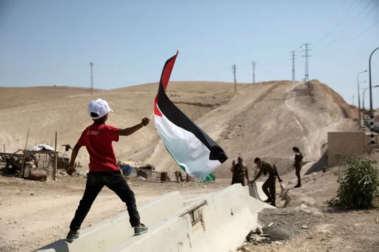 طفل فلسطيني يحمل علم فلسطين ويقف فوق حاجز، بينما يظهر جنود إسرائيليون في الخلفية، مما يعكس التوترات المستمرة في المنطقة.