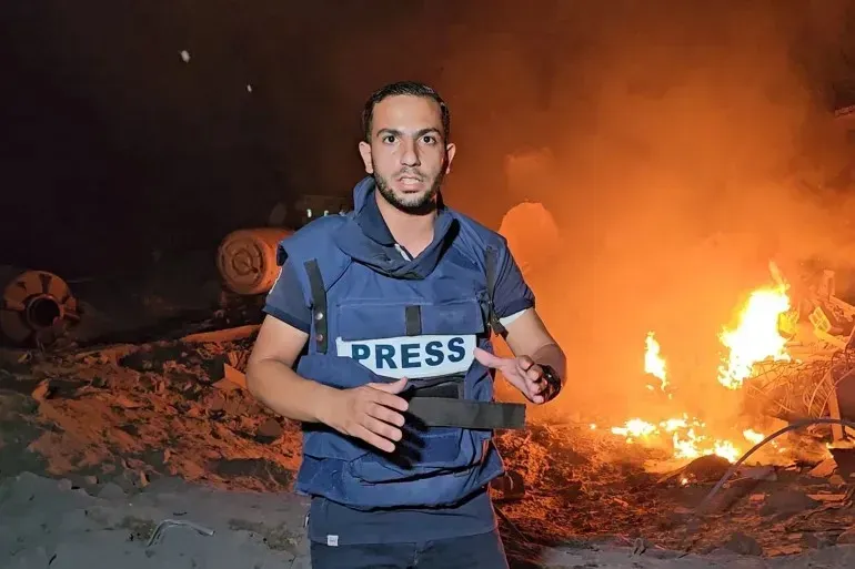صحفي يرتدي سترة "PRESS" يتحدث أمام حريق في منطقة متضررة بغزة، وسط أجواء من التوتر بعد القصف الإسرائيلي.