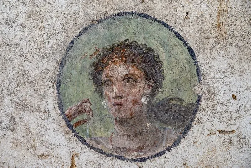 لوحة جدارية تُظهر امرأة ذات شعر مجعد تحمل شيئًا بيدها، تعكس الفنون الرومانية القديمة في بومبي، حيث عُثر على آثار مأساوية لضحايا ثوران بركان فيزوف.