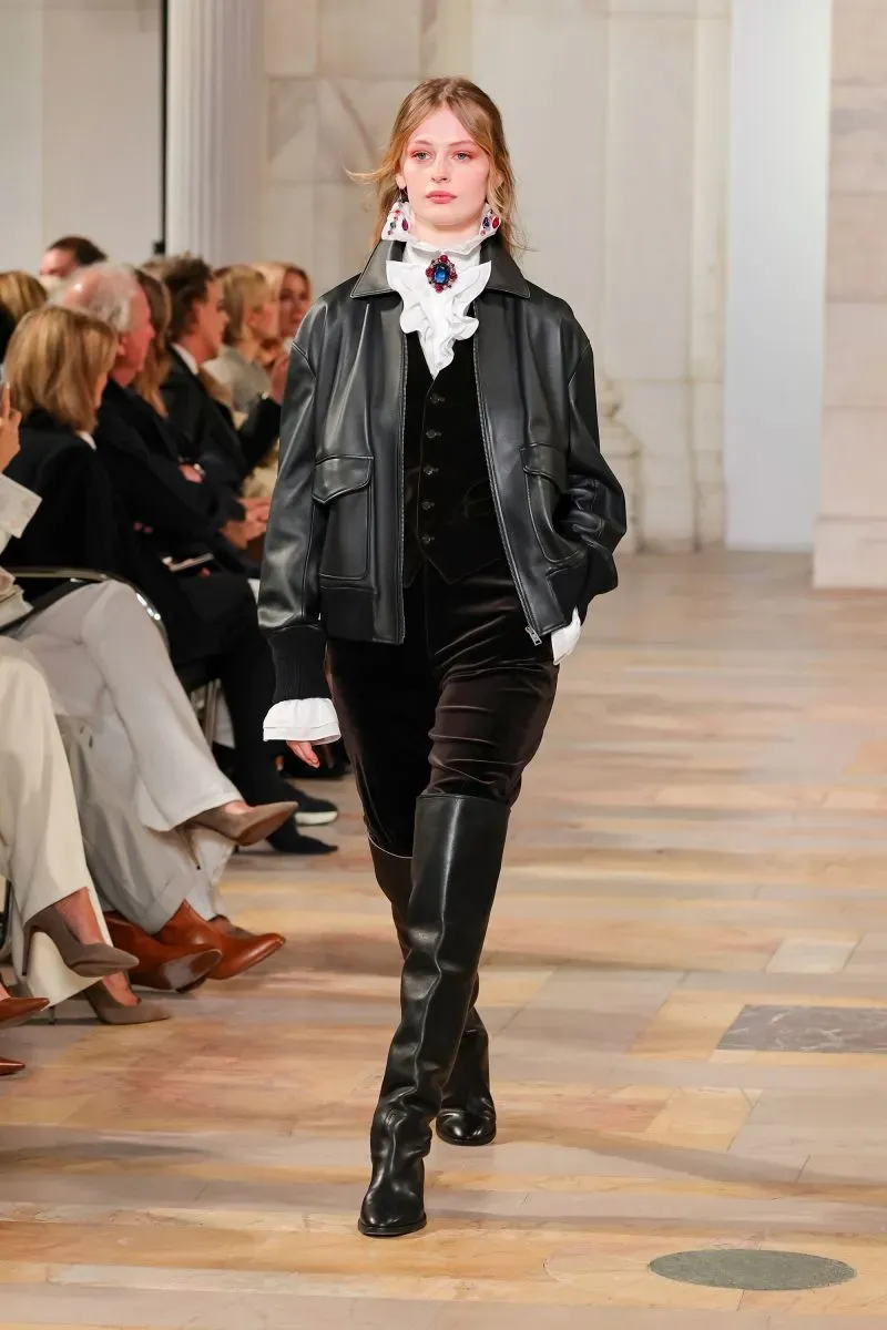 عارضة أزياء تسير على المنصة في عرض رالف لورين، ترتدي سترة جلدية سوداء وبنطلون مخملي، مع أجواء أنيقة تعكس مجموعة خريف 2025.