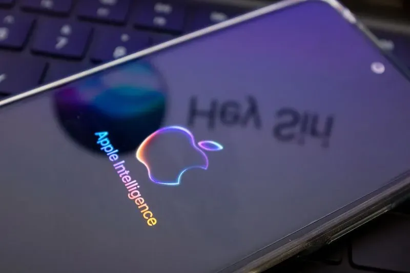 شعار "Apple Intelligence" يظهر على شاشة هاتف ذكي، مع انعكاس للوحة مفاتيح خلفية، مما يعكس تركيز آبل على الذكاء الاصطناعي.