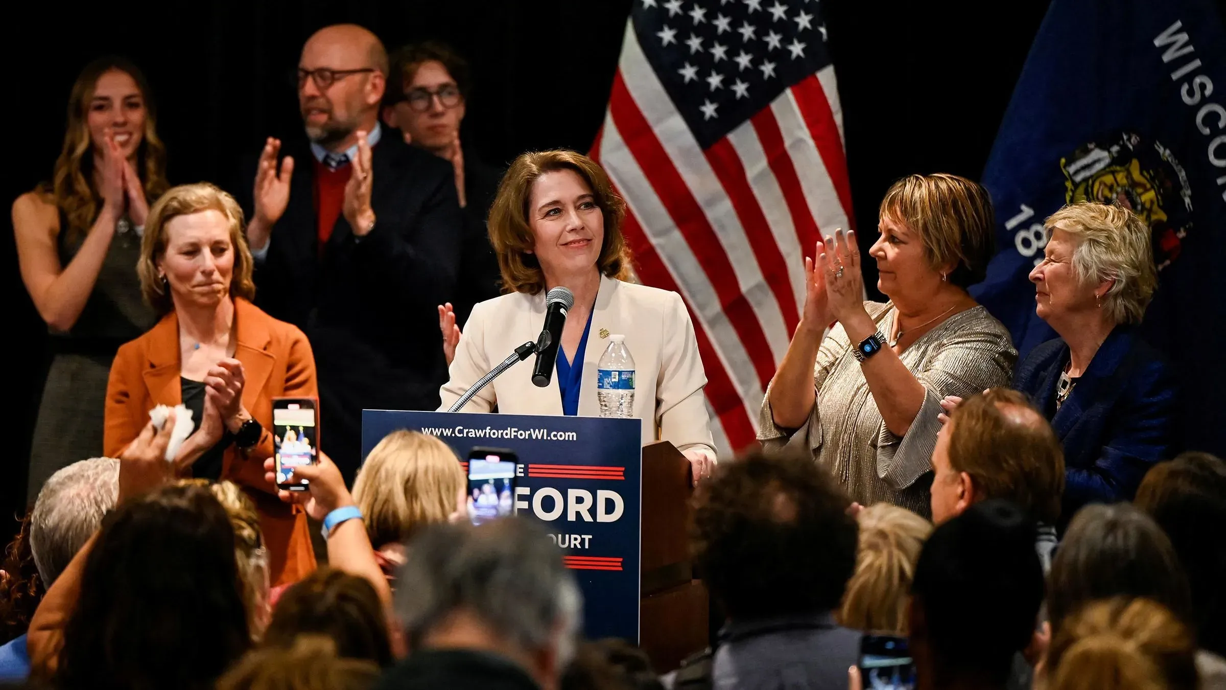 امرأة تتحدث على منصة انتخابية، محاطة بمؤيدين، مع العلم الأمريكي خلفها. تعكس الصورة أجواء الحماس خلال الانتخابات في ويسكونسن.