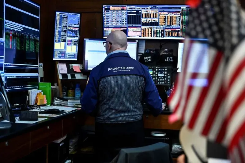 متداول في سوق الأسهم يقف أمام شاشات عرض متعددة تعرض بيانات مالية، مع علم أمريكي في الزاوية، يعكس التوترات الاقتصادية الحالية.