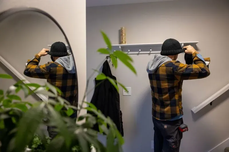 شخص يرتدي سترة مربعة وقبعة شتوية يقوم بتركيب رف في مدخل منزله، محاط بنباتات داخلية.