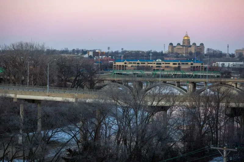 منظر لمدينة مع جسرين يمران فوق نهر، مع مبانٍ خلفية مضاءة بألوان غروب الشمس، مما يعكس التوترات العائلية في ظل السياسات الجديدة.