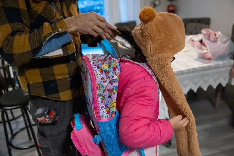 طفلة ترتدي سترة وردية وحقائب مدرسية ملونة، بينما يساعدها شخص بالغ في تسريح شعرها، في منزل عائلي دافئ.