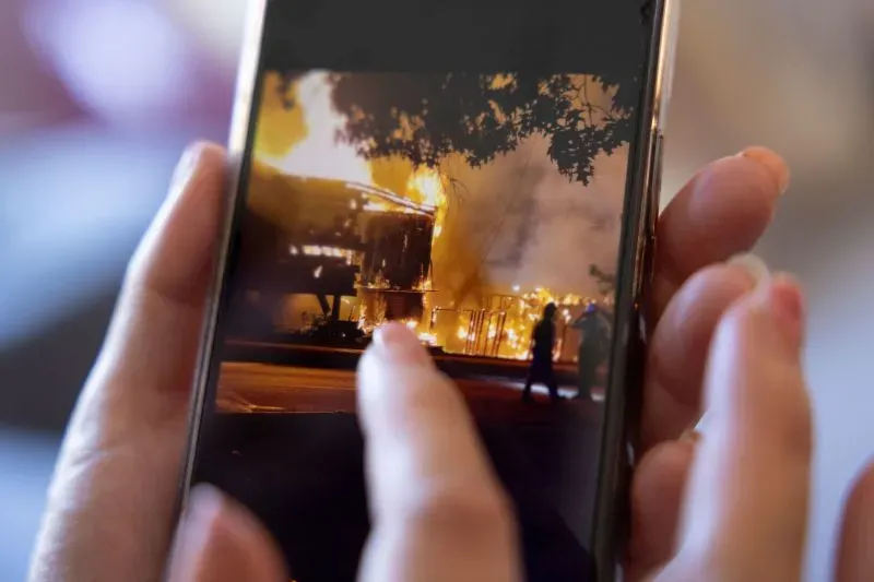 صورة تظهر يدًا تمسك بهاتف ذكي تعرض صورة لمنزل محترق، مع ألسنة اللهب والدخان، مما يعكس تجربة عائلة تواجه تحديات صعبة.