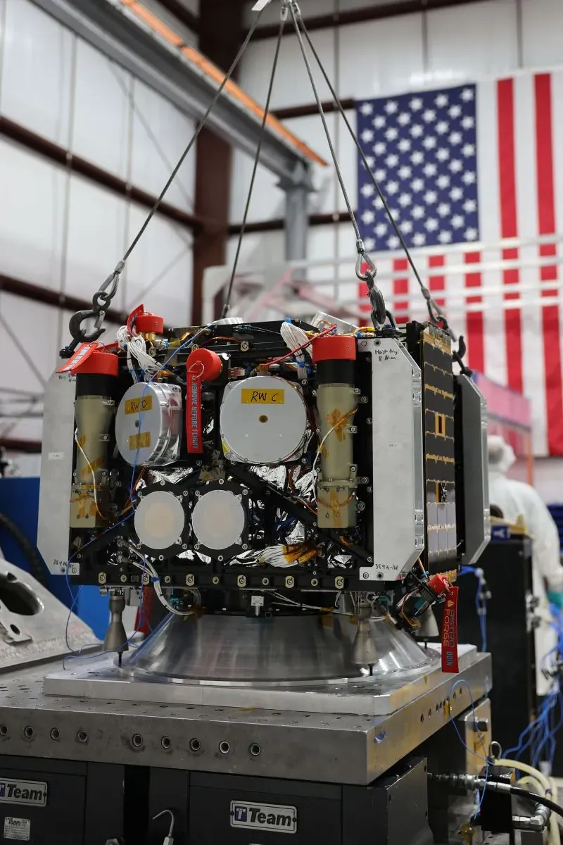 جهاز فضائي مُعلق داخل مختبر مع خلفية تتضمن علم الولايات المتحدة، يُظهر تفاصيل تكنولوجية معقدة.