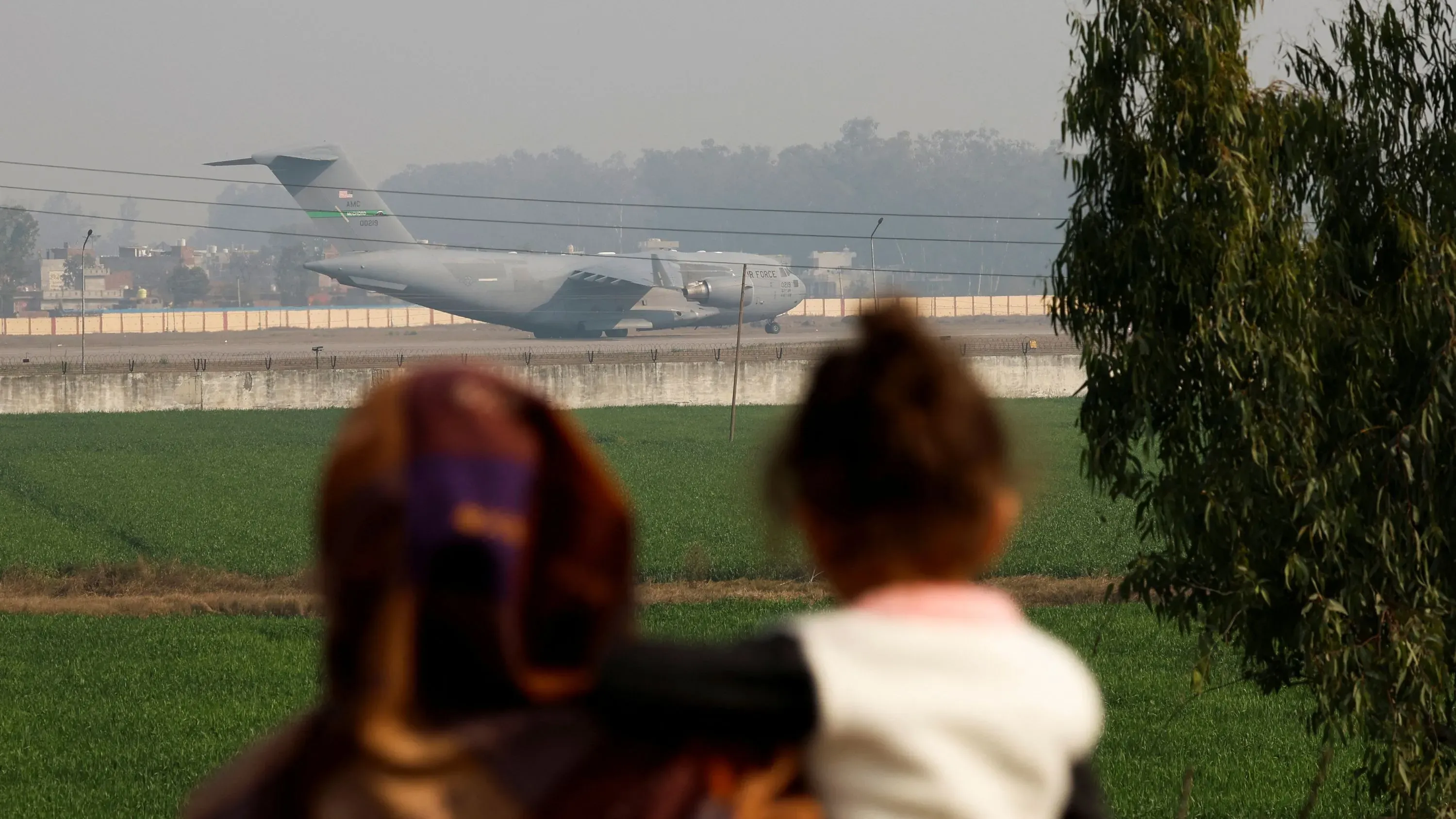طائرة عسكرية من طراز C-17 تهبط في أمريتسار بينما تراقب امرأة وطفلها، مع وجود حقول خضراء في الخلفية.
