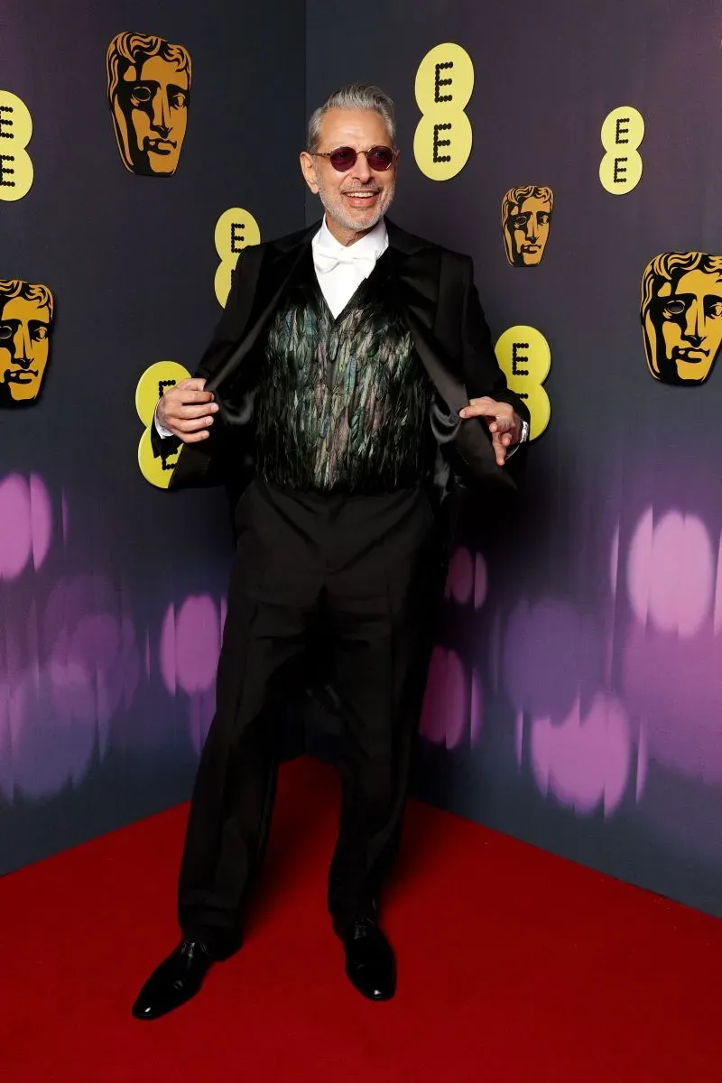جيف جولدبلوم مبتسمًا في حفل توزيع جوائز BAFTA، يرتدي بدلة أنيقة مع قميص أبيض ونظارات شمسية، خلفه خلفية تحمل شعار الجوائز.