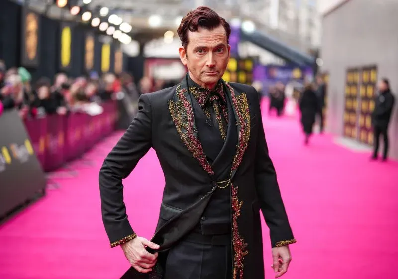ديفيد تينانت يرتدي بدلة أنيقة مزينة، يقف على السجادة الوردية في حفل توزيع جوائز BAFTA، مع حشد من المعجبين في الخلفية.