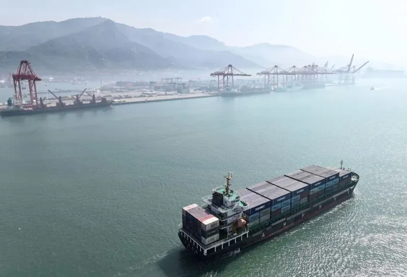 سفينة شحن تمر عبر ميناء مزدحم بالصين، مع وجود حاويات وشاحنات على الأرصفة، مما يعكس النشاط التجاري المتزايد وسط التوترات التجارية.