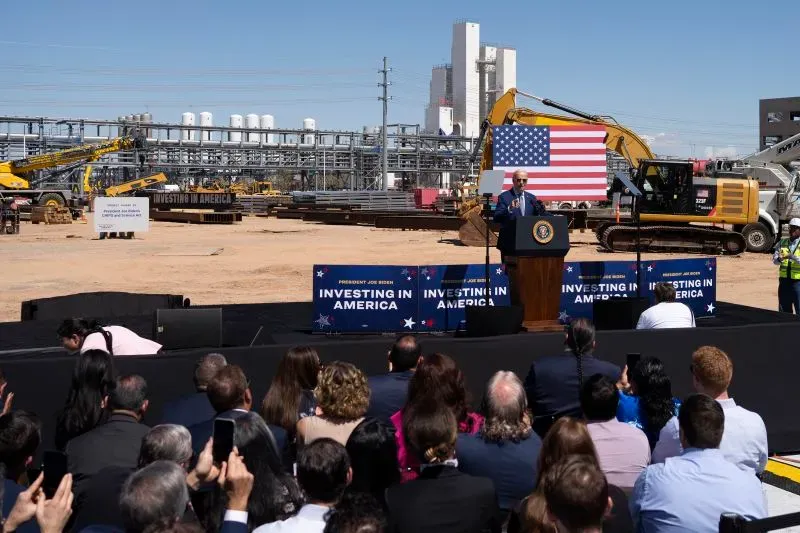 بايدن يلقي خطابًا أمام جمهور خلال حدث للاستثمار في أمريكا، مع معدات بناء وخلفية العلم الأمريكي.