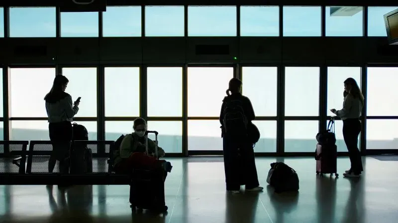 مسافرون ينتظرون في مطار، حيث تظهر silhouettes لأشخاص يحملون حقائبهم ويتواصلون عبر الهواتف، مع خلفية نوافذ مضاءة.